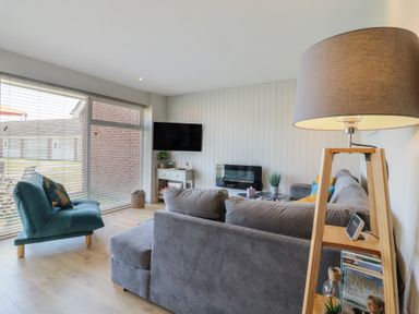 A living room with a grey sofa a teal chair a floor lamp and a TV mounted on the wall at 1 Kessingland Cottages in Kessingland