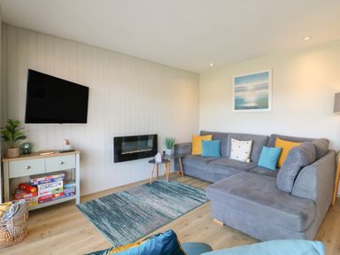 A living room with a gray sectional sofa with colorful cushions a wall mounted television wooden floor and a small console table with board games at 1 Kessingland Cottages in Kessingland