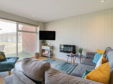 A living room with a gray sofa and colorful cushions a wall-mounted tv and a small table holding games at 1 Kessingland Cottages in Kessingland