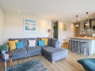 A living room with a gray sectional sofa and colorful cushions near a kitchen island with three stools at 1 Kessingland Cottages in Kessingland