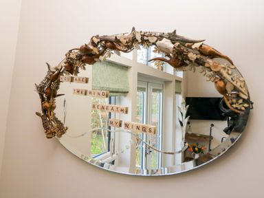 An oval mirror with a decorative natural wood frame and letter tiles forming the phrase you are the wind beneath my wings reflected in the mirror at Elderflower Lodge in Allithwaite near Cartmel