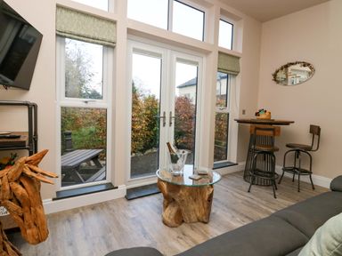 A living area with glass doors overlooking a garden a glass and wood coffee table a wooden round table with two chairs and a TV mounted on the wall at Elderflower Lodge in Allithwaite near Cartmel