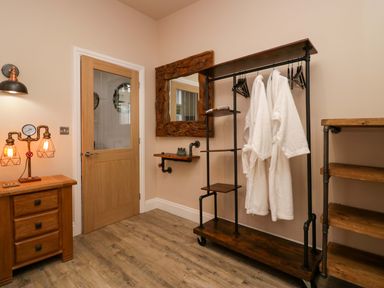 A room with wooden furniture two white bathrobes hanging on a metal and wood rack a wooden door a mirror with a wooden frame and a wall lamp at Elderflower Lodge in Allithwaite near Cartmel