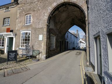 A stone archway over a narrow street with old buildings on either side and a bench with metal sculptures outside a shop