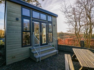 A small wooden building with glass doors and windows next to a picnic table outdoors at Elderflower Lodge in Allithwaite near Cartmel