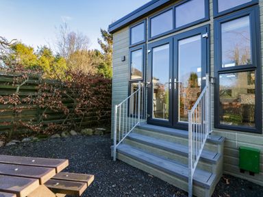 An exterior view of a building with glass doors and steps leading to them a wooden fence and trees in the background with a wooden picnic table in the foreground at Elderflower Lodge Allithwaite near Cartmel
