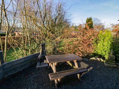 A wooden picnic table with benches on gravel near trees shrubs and a small outdoor fireplace at Elderflower Lodge in Allithwaite near Cartmel