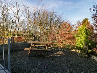 An outdoor seating area with a wooden picnic table surrounded by trees and shrubs at Elderflower Lodge in Allithwaite near Cartmel
