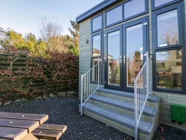 Exterior view of a building with steps leading to glass doors and a gravel ground with a wooden picnic table nearby at Elderflower Lodge in Allithwaite near Cartmel