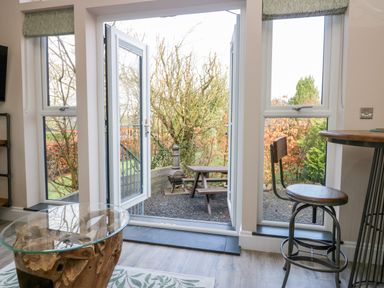 A room with glass doors leading to an outdoor area with a wooden picnic table and metal stove at Elderflower Lodge in Allithwaite near Cartmel