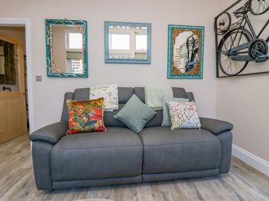 A gray sofa with five patterned cushions and two blankets in a room with three mirrors and a bicycle wall art at Elderflower Lodge in Allithwaite near Cartmel