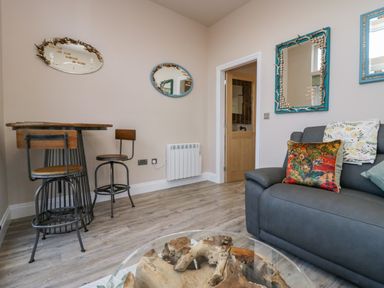 A living room with a gray sofa with cushions a round glass coffee table wooden chairs and a tall table wooden door and mirrors on the wall at Elderflower Lodge in Allithwaite near Cartmel