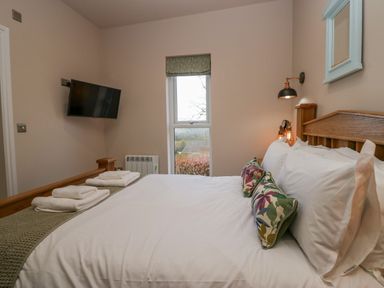 A bedroom with a wooden bed with white bedding and floral pillows a wall-mounted television and a window at Elderflower Lodge in Allithwaite near Cartmel