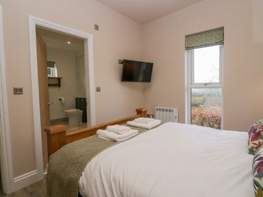 A bedroom with a wooden bed a wall mounted tv towels on the bed and an en suite bathroom at Elderflower Lodge in Allithwaite near Cartmel