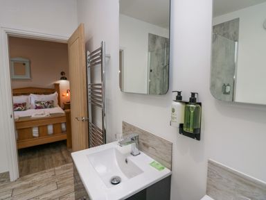 A bathroom sink with soap dispensers and mirrors showing a bedroom with a wooden bed at Elderflower Lodge in Allithwaite near Cartmel