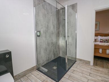 A bathroom with a glass shower enclosure next to a black toilet and a view of a bedroom with a wooden bed frame at Elderflower Lodge in Allithwaite near Cartmel