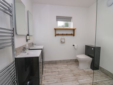 A bathroom with two sinks with mirrors above them a toilet a wooden shelf with hooks and a heated towel rail at Elderflower Lodge in Allithwaite near Cartmel