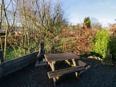 An outdoor seating area with a wooden picnic table bench and a chiminea surrounded by trees and shrubs at Elderflower Lodge in Allithwaite near Cartmel