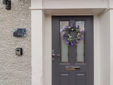 A front door with two small windows and a wreath with purple flowers next to a wall plaque reading 1 Brookside in Warcop