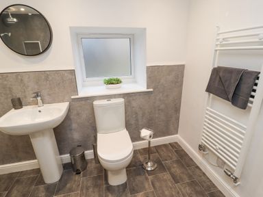 A bathroom with a pedestal sink and round mirror a toilet and freestanding toilet paper holder a towel rack with towels and a frosted window with a plant on the sill at 1 Brookside in Warcop