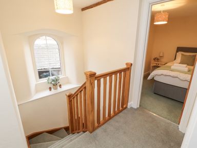 A hallway with wooden staircase railing a window with flowers on the sill and a bedroom with a bed and bedside table at 1 Brookside in Warcop