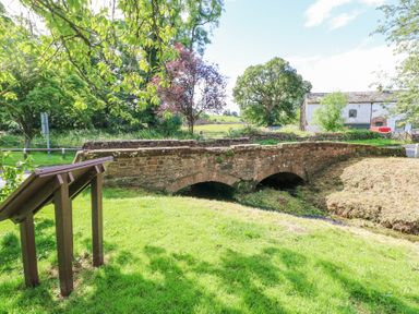 A stone bridge over a small stream with a wooden information board and trees at 1 Brookside in Warcop