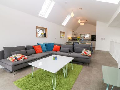 A living area with a gray sectional sofa with colorful pillows and blankets a white coffee table on a green rug and a dining area in the background at Ty Chwarel in Menai Bridge