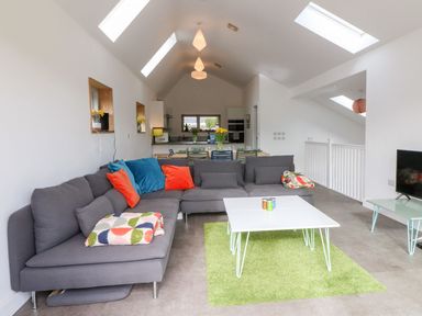 An open living room and kitchen with a gray sectional sofa with colorful cushions and blankets a white coffee table on a green rug a dining table with chairs and flowers and skylights on the ceiling at Ty Chwarel in Menai Bridge