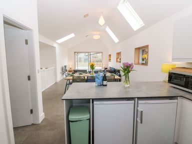 A kitchen area with a countertop and two small refrigerators next to a dining table with flowers in vases and a living area in the background at Ty Chwarel in Menai Bridge