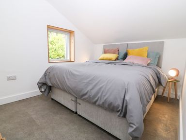A bedroom with a bed covered in grey bedding and colorful pillows next to a window and bedside table with a lamp at Ty Chwarel in Menai Bridge