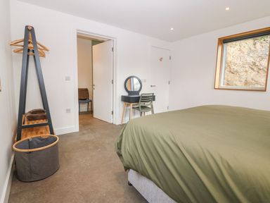 A bedroom with a bed covered in green bedding a window a chair a small desk with a mirror an open door a clothes rack and a laundry basket at Ty Chwarel in Menai Bridge
