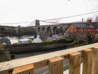 A view of a stone suspension bridge over a river with houses and a wooden railing in the foreground at Ty Chwarel in Menai Bridge