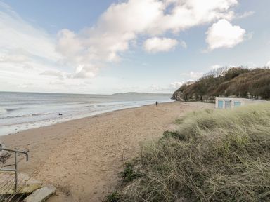A sandy beach with a person walking near a rocky outcrop and a small building with blue doors by the shore at Seaview in Benllech near Red Wharf Bay