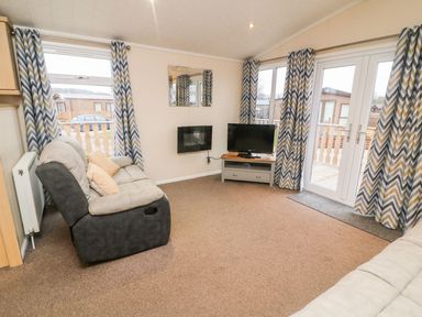 A living room with two sofas a wall mounted fireplace a TV on a stand and windows with patterned curtains at 6 Sherwood in Carnforth near Tewitfield