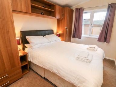 A bedroom with a double bed white bedding folded towels bedside tables with lamps wooden wardrobes and a window with curtains at 6 Sherwood Carnforth near Tewitfield