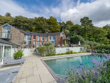An outdoor swimming pool with lounge chairs and a stone house surrounded by trees at Curlew House in Cosheston