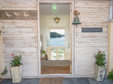 An open doorway with wooden panel walls and flowerpots on each side overlooking a water view at Curlew House in Cosheston