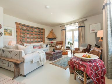 A living room with sofas, armchairs, a coffee table, a wooden bench, a model sailboat, and a table with a red cloth and fruit bowl at Curlew House in Cosheston