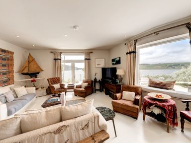 A living room with leather chairs white sofa model sailboat and large windows overlooking water at Curlew House in Cosheston