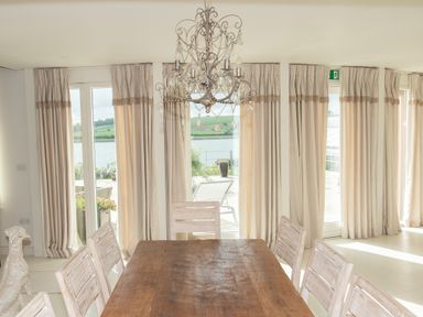 A dining area with a wooden table and chairs under a chandelier with windows and curtains showing an outdoor view at Curlew House in Cosheston