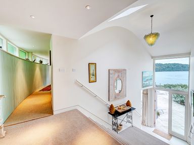 A hallway with a staircase leading down to glass doors overlooking a body of water with a curved green wall and art on the walls at Curlew House in Cosheston