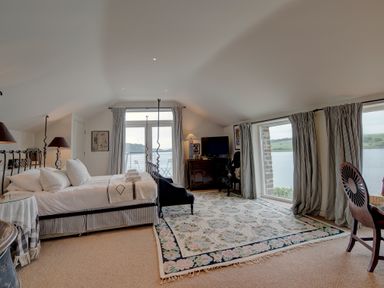 A bedroom with a bed pillows a floral rug lamp and television overlooking water at Curlew House in Cosheston