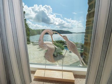 Two wooden bird sculptures on a windowsill with striped curtains and a view of a patio and water beyond at Curlew House in Cosheston