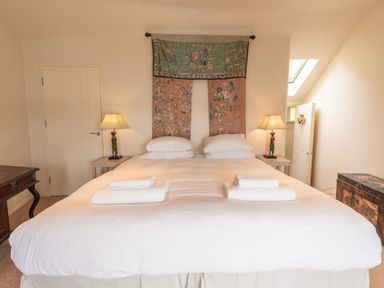 A bedroom with a large bed with white bedding and folded towels two bedside tables with lamps and decorative figures a tapestry hanging on the wall and an antique chest at Curlew House in Cosheston