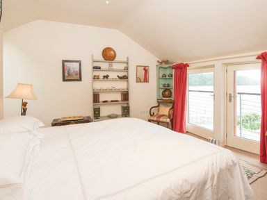 A bedroom with a bed lamp bookshelf armchair and glass doors with red curtains at Curlew House in Cosheston