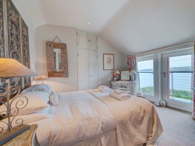 A bedroom with a bed white bedding wooden mirror lamps and glass doors overlooking water at Curlew House in Cosheston