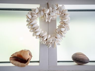 A window with a heart-shaped shell wreath hanging in the center and two large seashells on the sill at Curlew House in Cosheston
