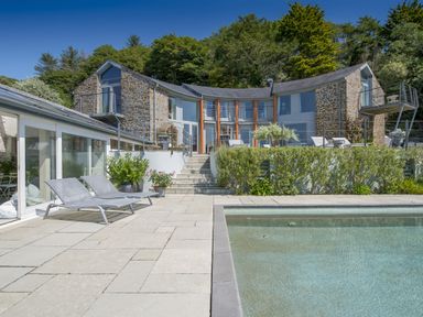 A stone house with large windows and a curved design next to a swimming pool and lounge chairs at Curlew House in Cosheston