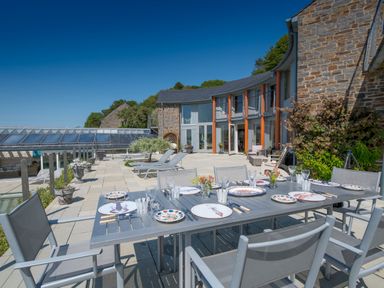 An outdoor patio with a dining table set with plates and glasses near a stone building with large windows at Curlew House in Cosheston