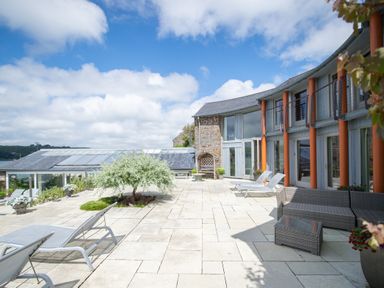 An outdoor patio with lounge chairs a small sofa set a stone planter with a tree and a building with large windows at Curlew House in Cosheston